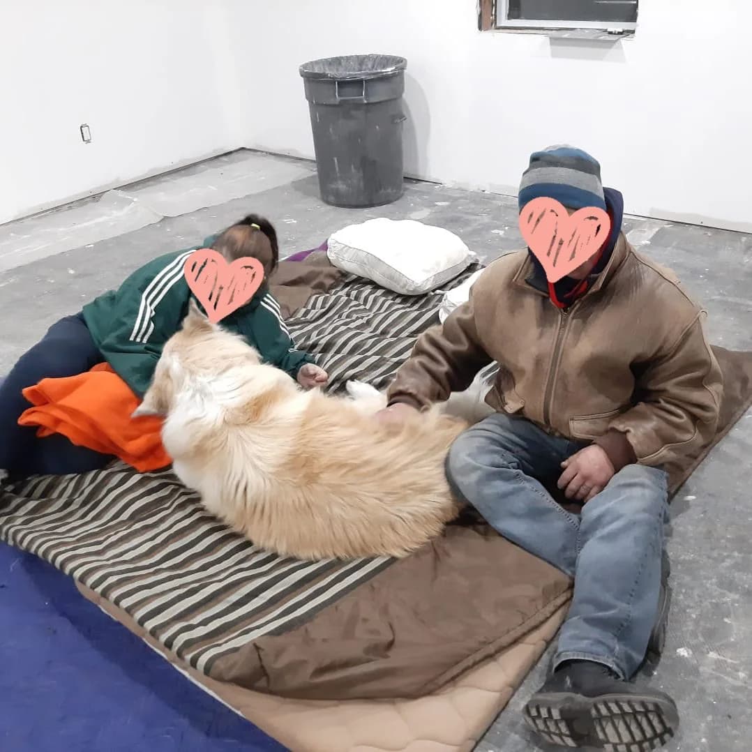Buddy resting with two people inside the temporary warming space at the BuildX shop. Faces are covered.